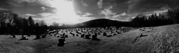 wide, black and white view of a cemetery on a grassy hillside with scattered tombstones, bare trees, and a bright sun low in the sky.