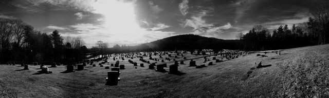 wide, black and white view of a cemetery on a grassy hillside with scattered tombstones, bare trees, and a bright sun low in the sky.