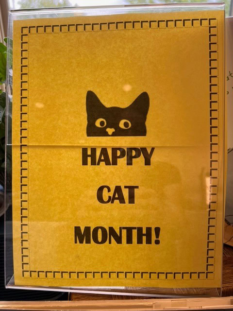 a yellow sign with a black cat graphic and the text "happy cat month!" in bold black letters.