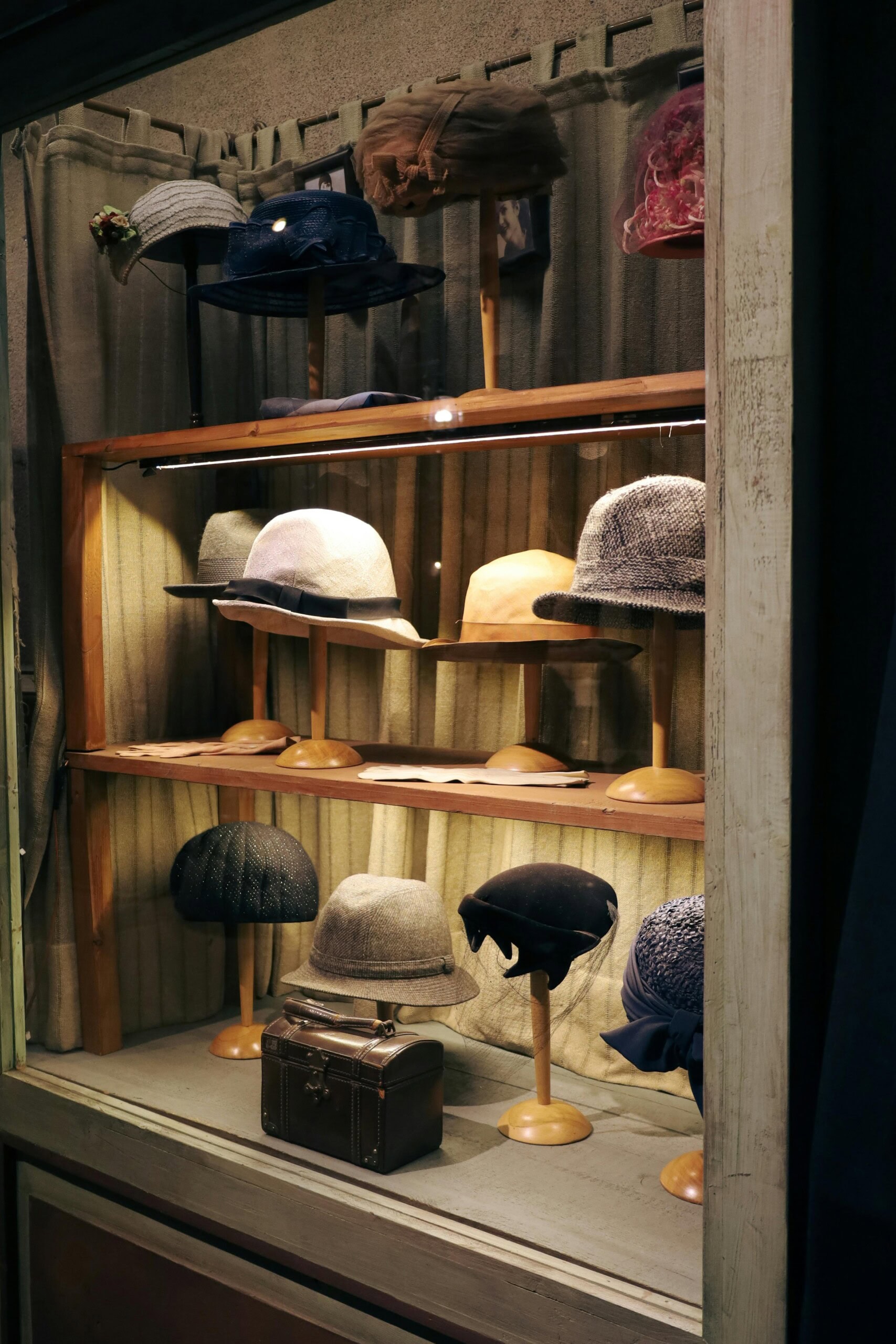 a display case with shelves holding various vintage hats on stands and a small brown suitcase at the bottom.