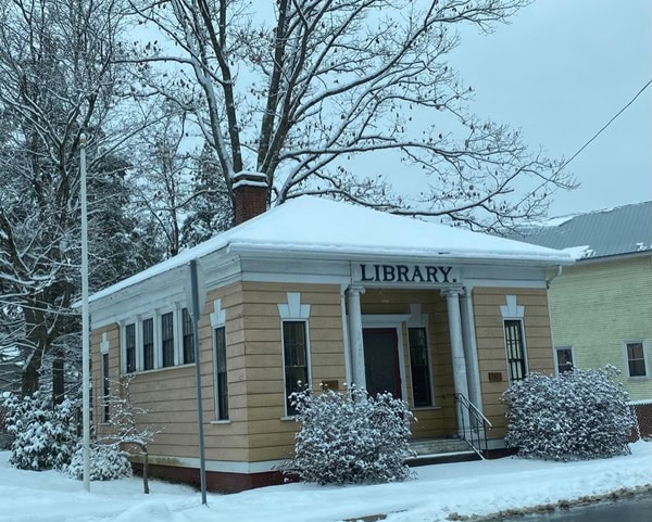 a small, single story building with columns and a sign reading "library" on the front, surrounded by snow covered trees and bushes.
