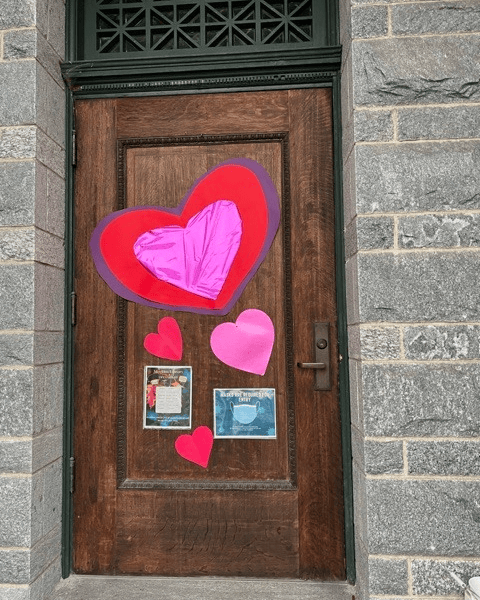a wooden door decorated with large and small paper hearts in red and pink, and two printed flyers taped near the bottom.