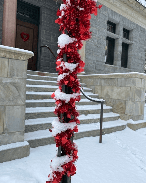 a metal pole wrapped with red tinsel and snow stands in front of a stone building with steps and a heart decoration on the door. snow covers the ground and steps.