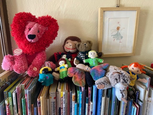 stuffed animals and dolls are arranged on top of a bookshelf filled with children's books, with a framed fairy illustration hanging on the wall above.