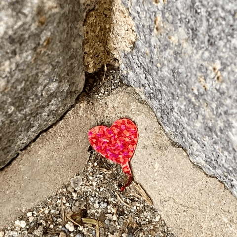 a small, red glitter heart sticker lies on the ground between two rough concrete blocks and some dirt.