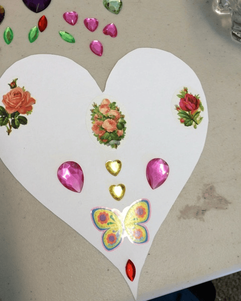 a white paper heart decorated with floral stickers, gem stickers in pink, yellow, red, and green, and a colorful butterfly sticker on a gray surface.