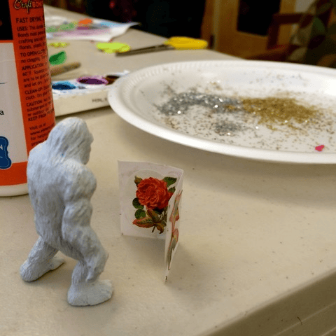 a small yeti figurine stands in front of a handmade card with a rose, next to a paper plate with glitter and craft supplies.