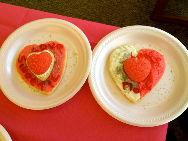 two heart shaped cookies on white plates, decorated with red and white icing, red sugar hearts, and silver sprinkles, placed on a pink tablecloth.