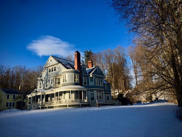 large blue victorian style house with a wraparound porch sits on a snowy lawn, surrounded by leafless trees under a clear blue sky.