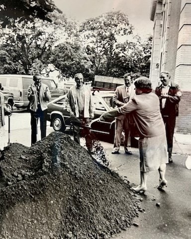 a woman shovels dirt from a pile while several men stand nearby watching outside next to parked cars and a building.