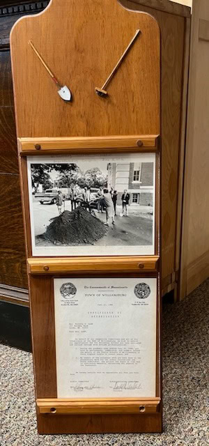 a wooden display holds two small gardening tools, a black and white photo of people shoveling dirt, and a framed official document with typed and handwritten text.