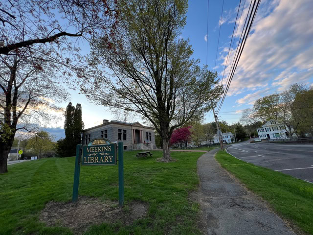a sign reading “meekins library” stands on a grassy area beside a tree lined sidewalk, with the library building and houses visible under a partly cloudy sky.