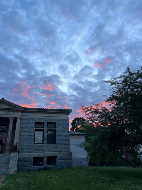 stone building and trees in the foreground with a cloudy sky tinged with pink from the setting sun in the background.
