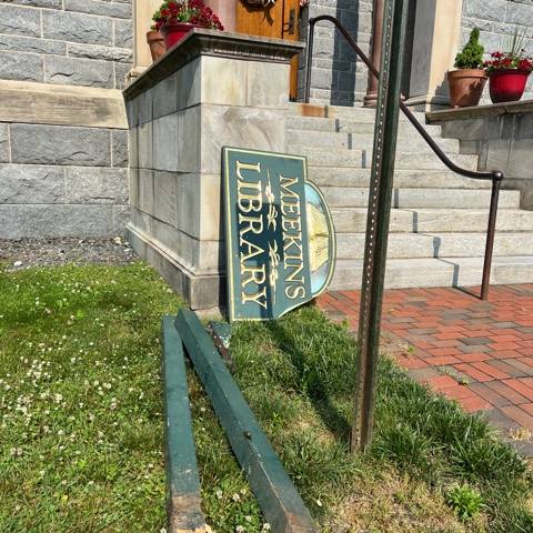 a library sign reading "meekins library" lies on the ground next to two broken posts in front of a stone building with steps and potted plants.