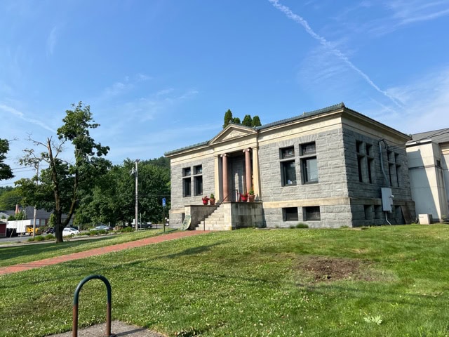 stone public building with columns, wide steps, and tall windows, set on a grassy lawn with a clear blue sky and a few trees in the background.