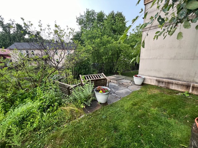 wooden outdoor bench and potted plants on a stone patio next to a building, surrounded by grass, greenery, and a black metal fence.