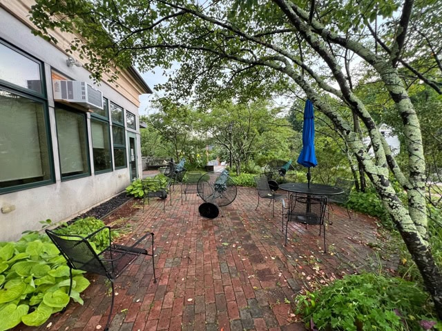 brick patio with several black metal chairs and tables, some toppled over, surrounded by green plants and trees next to a building with large windows.
