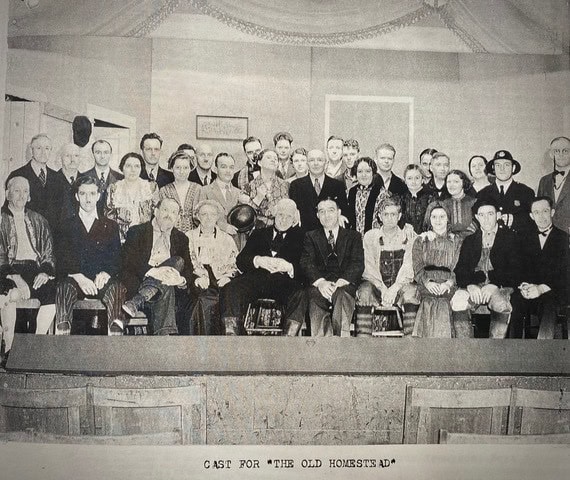 a large group of men and women in period costumes pose together on stage for a cast photo labeled 