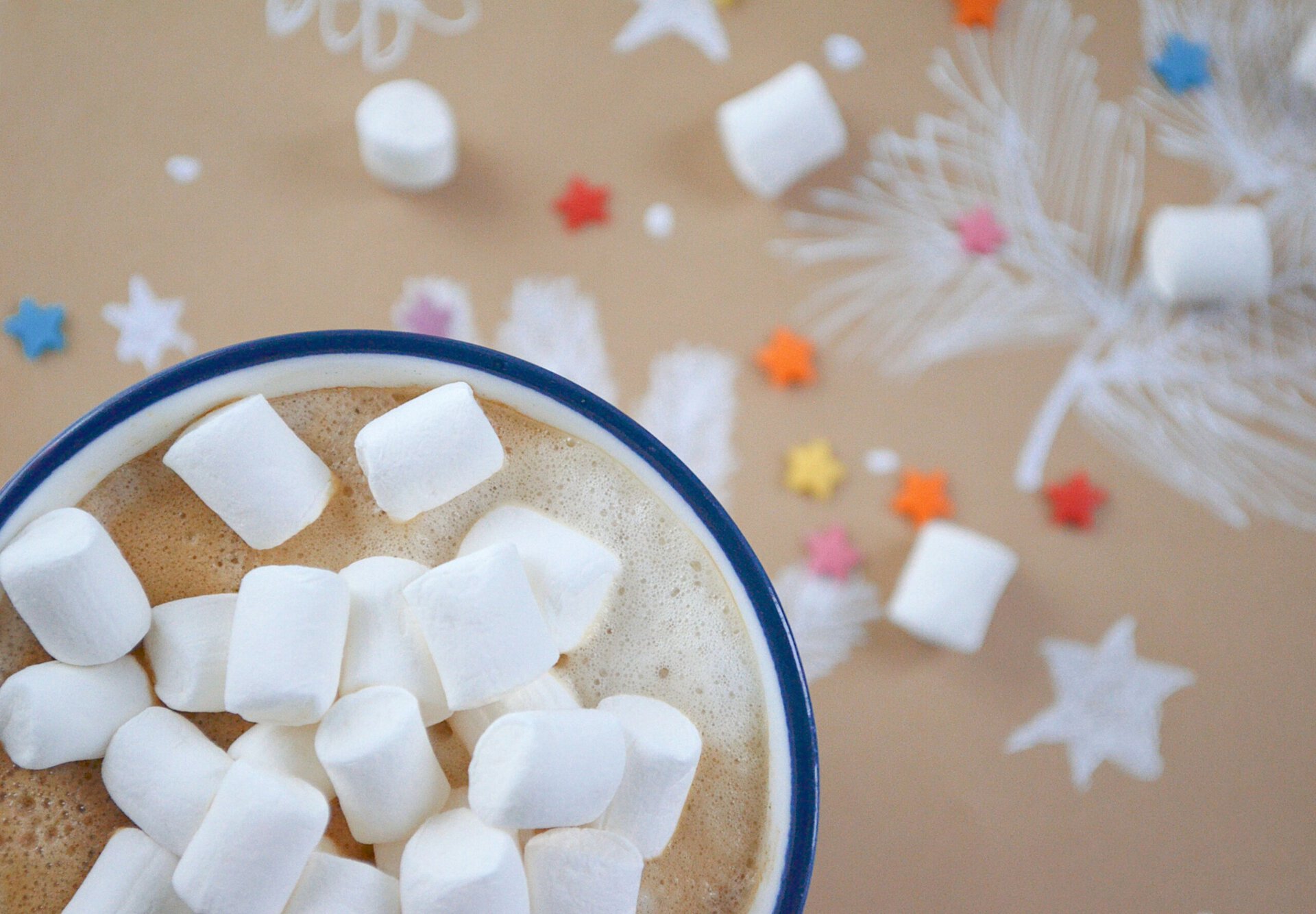 a mug of hot cocoa topped with marshmallows, set against a brown background decorated with white drawings of fir branches and colorful star shaped sprinkles.