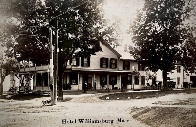 black and white photo of hotel williamsburg in massachusetts, featuring a two story building with a covered porch, large trees, and several people and horse drawn carriages outside.