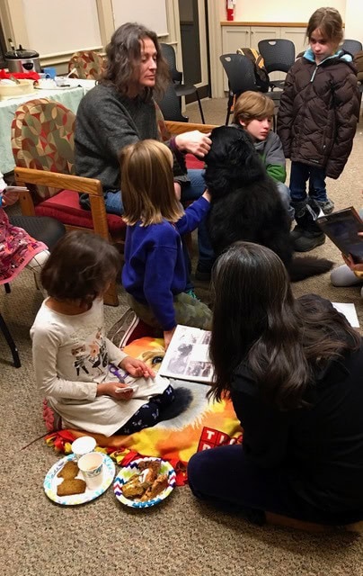 a group of children and adults sit and stand around a large black dog indoors. some children are petting the dog, while others are drawing or eating snacks.
