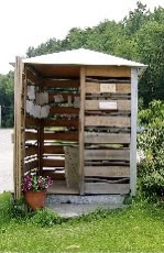 a small wooden shelter with slatted walls and a metal roof stands on grass, with a potted flower placed at the entrance. trees and greenery are visible in the background.
