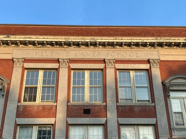facade of a brick building with "1914 helen e. james school 1914" engraved above two sets of large windows, lit by late afternoon sunlight.