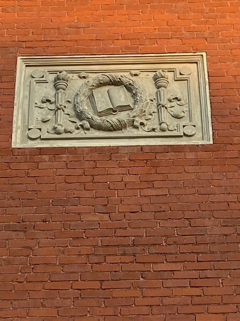 a stone relief of an open book surrounded by decorative elements is set into a red brick wall.