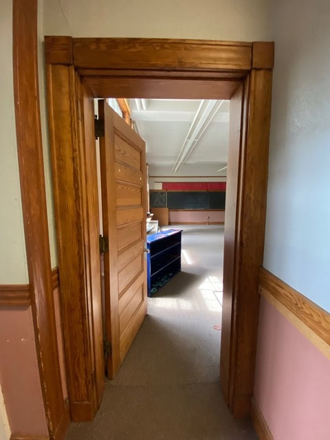 a doorway with wooden trim opens into a room with a large window, shelves, and blackboards on the far wall. the floor is carpeted, and the walls have wood paneling.