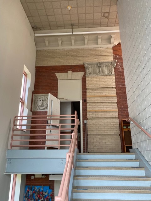 a stairwell with metal railings leads to a landing with an open door; exposed brick, decorative stonework, and a high ceiling are visible on the walls.