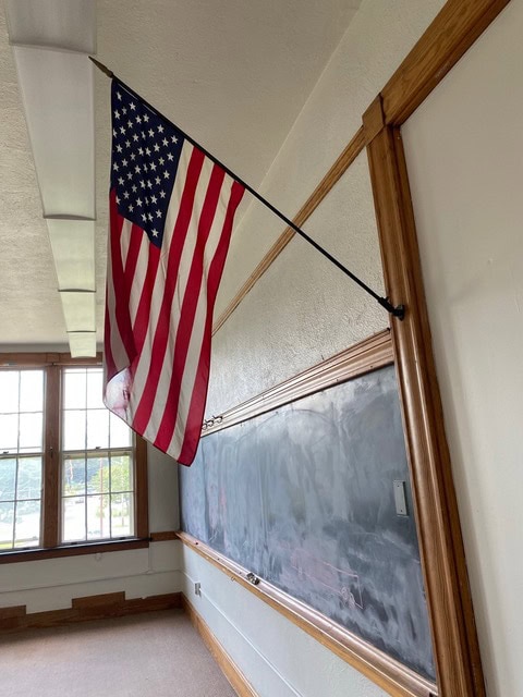an american flag hangs on a wall above a chalkboard in a classroom with large windows and natural light.