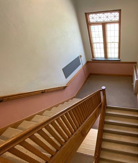 an indoor stairwell with wooden railings and beige steps leads down to a landing near a large window with multiple panes.