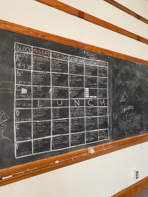 a classroom chalkboard displays a weekly schedule grid with colored chalk for each day and time block, featuring "lunch" written diagonally across the middle blocks.
