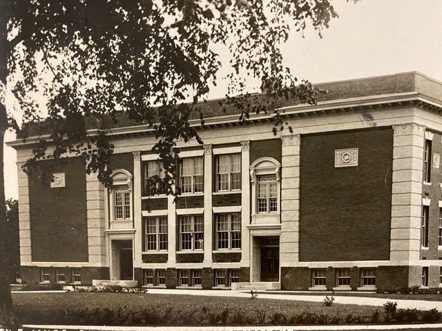 a two story brick building with large windows and decorative trim, surrounded by grass and partially framed by tree branches.