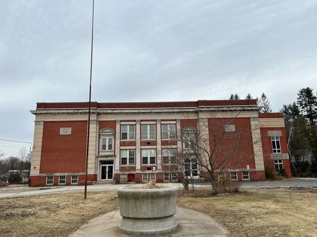 a two story red brick building with white stone trim, large windows, and an empty flagpole stands behind a round concrete structure on a cloudy day.