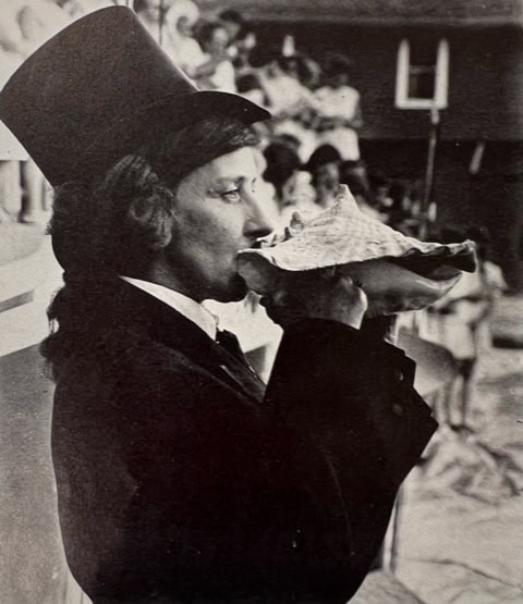 person in a suit and top hat blows into a large conch shell, with a blurred crowd in the background.