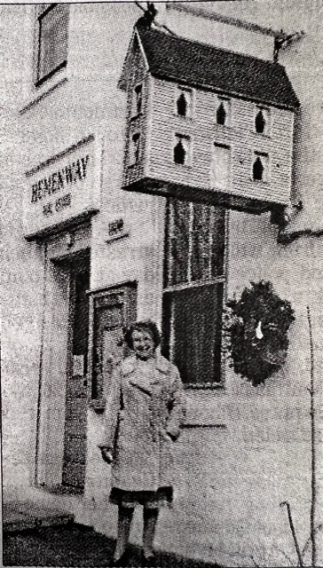 a woman stands outside a building labeled "hemenway," beneath a large decorative birdhouse mounted above the entrance.