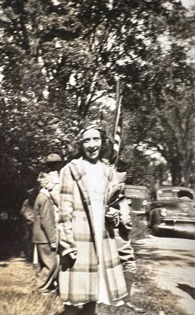 a young woman in a plaid coat stands outdoors, holding items in her arms, with people, trees, and vintage cars visible in the background.