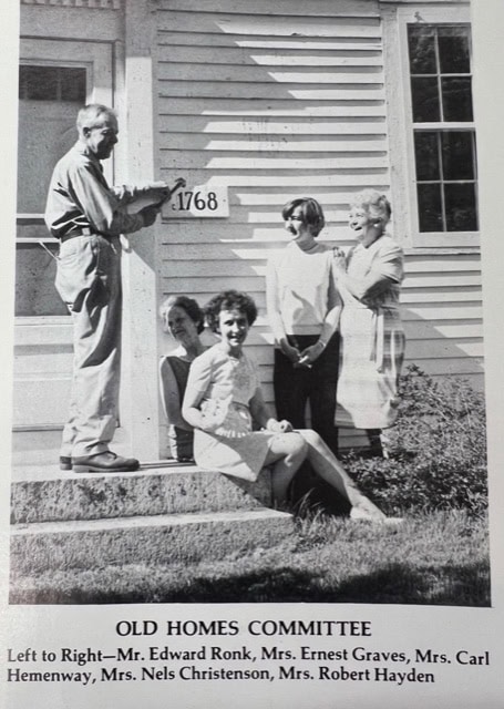five adults stand and sit on the steps of a house labeled "1768," with a sign reading "old homes committee" and their names listed below the photo.