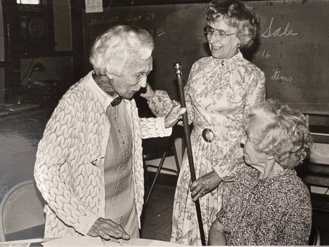 three elderly women are talking and smiling together in a classroom. one stands with a cane, another stands beside her, and the third woman is seated. a chalkboard is visible in the background.