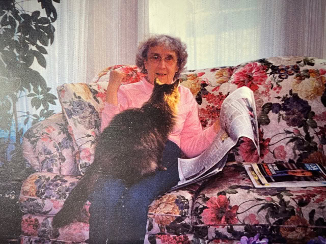 an elderly woman sits on a floral couch reading a newspaper while a long haired cat stands on her lap, nuzzling her face.