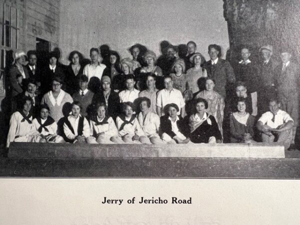a black and white group photo of men, women, and children posing in rows; caption reads 