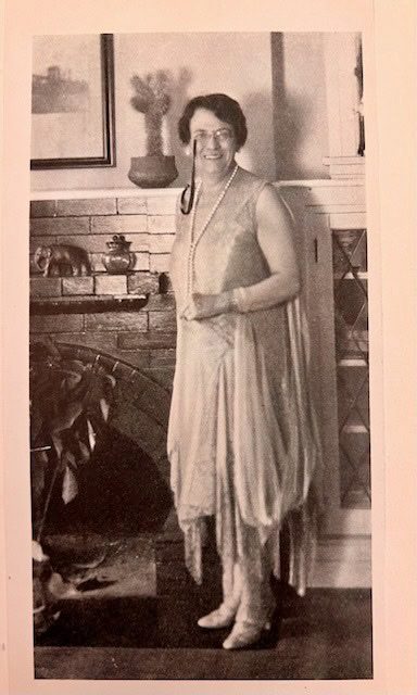 a woman wearing a sleeveless, knee length dress and long necklace stands in front of a brick fireplace, smiling at the camera.
