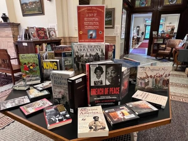 a library display table features books and dvds about african american history, civil rights, and juneteenth, set in a reading area with bookshelves and a decorative tiled floor.