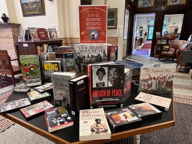 a library display table features books and dvds about african american history, civil rights, and juneteenth, set in a reading area with bookshelves and a decorative tiled floor.
