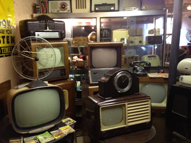 a collection of vintage radios, televisions, a rotary phone, and a clock are displayed on shelves and tables in a room.