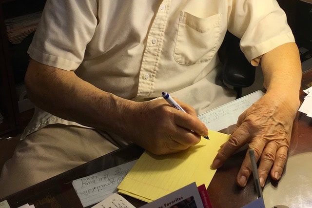an older person in a short sleeve white shirt writes on a yellow notepad at a desk cluttered with papers and notes.