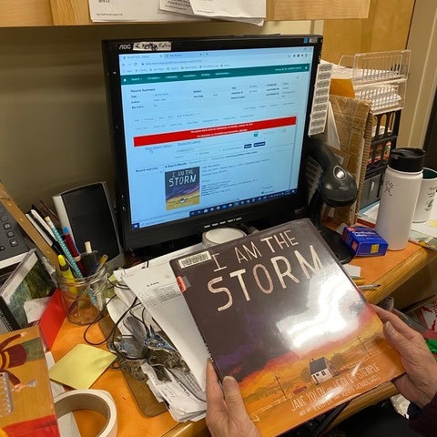 person holding the book "i am the storm" at a cluttered desk with a computer displaying information about the same book.