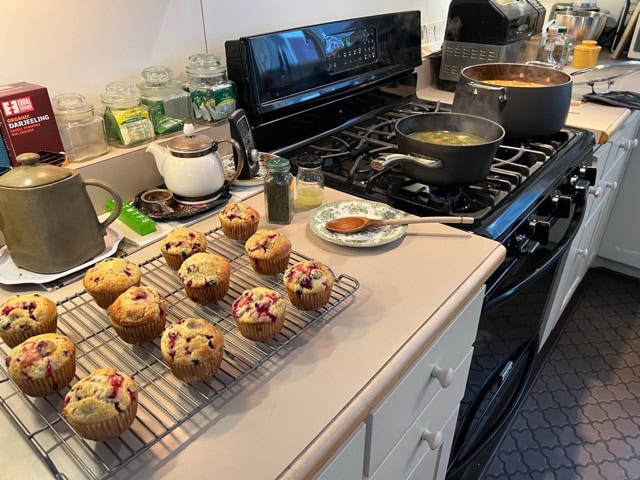 a kitchen scene with muffins cooling on a rack, pots simmering on the stove, and various kitchen items on the counter.