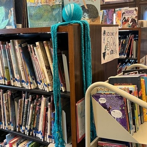 a ball of turquoise yarn with long strands drapes over the top corner of a library bookshelf filled with books. a book cart is nearby.